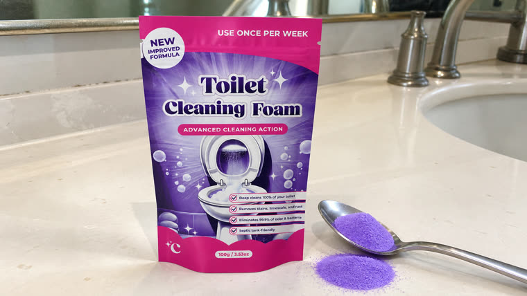 The pink FizzClean Toilet Cleaning Foam pouch standing on a bathroom counter beside a tablespoon heaped with violet powder